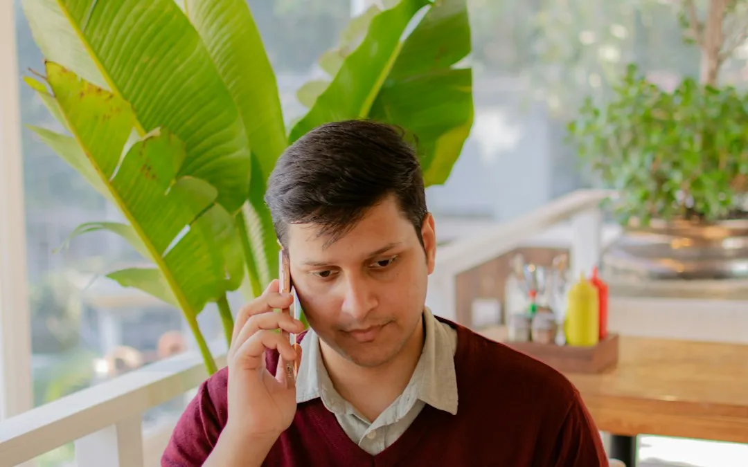 A man at a table by a window, listening on a phone call.