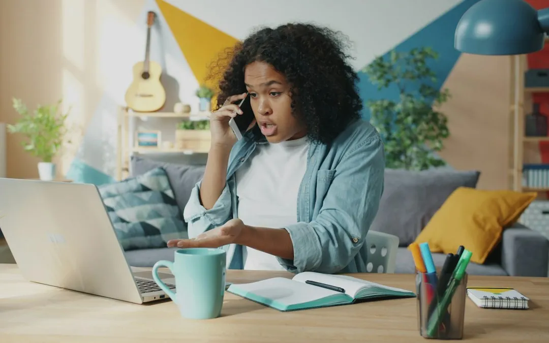A woman at a desk with a laptop and notebook, talking through something on the phone.